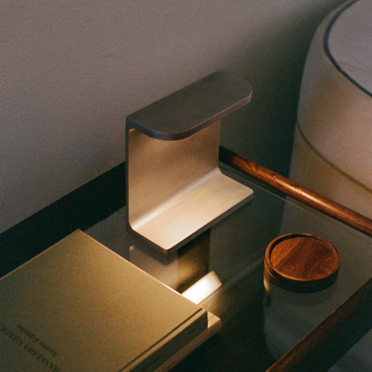 A lit aluminium desk lamp on a glass side table beside a book and round wooden coaster